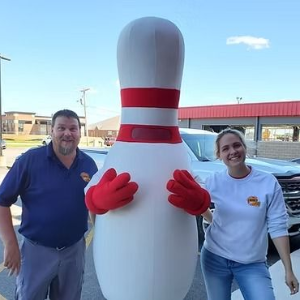 Two people posing with a giant bowling pin mascot outside a bowling alley