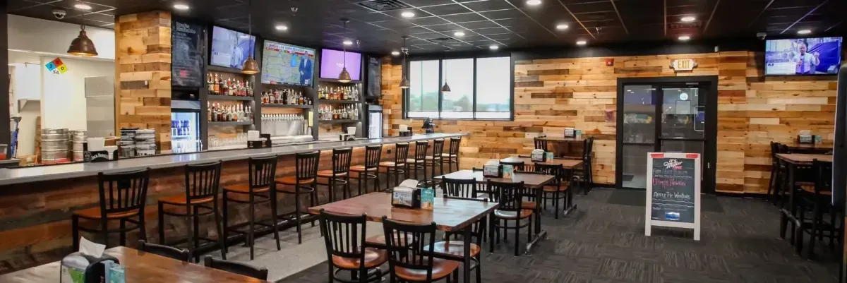 Empty sports bar with wooden tables, stools, TVs, and a menu board under warm lighting.