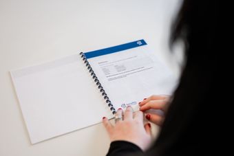Person holding open spiral-bound notebook with text on a white table.