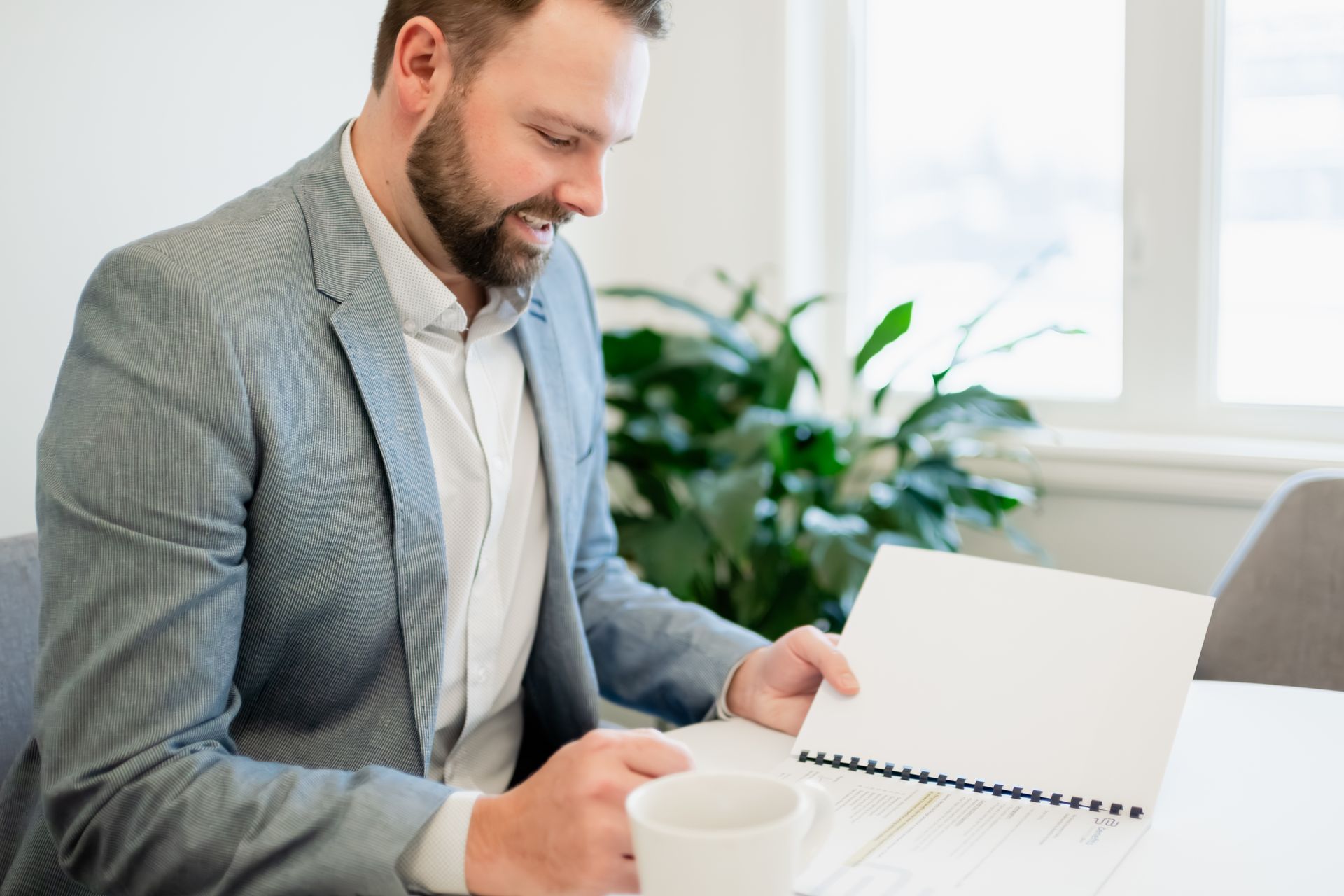Man in a gray blazer looks at paperwork with a smile, coffee mug on the table.