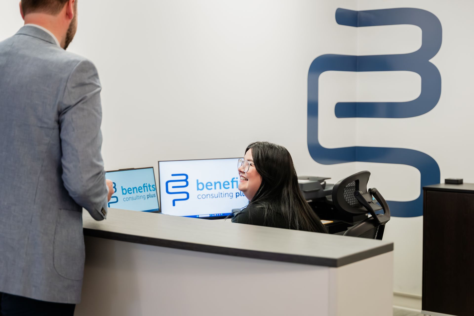 Person at a reception desk smiles at someone approaching. The company logo is visible.