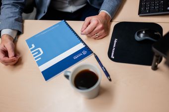 Person at desk with notebook, pen, coffee, mouse, and keyboard. Blue and white notebook, light tan desk.