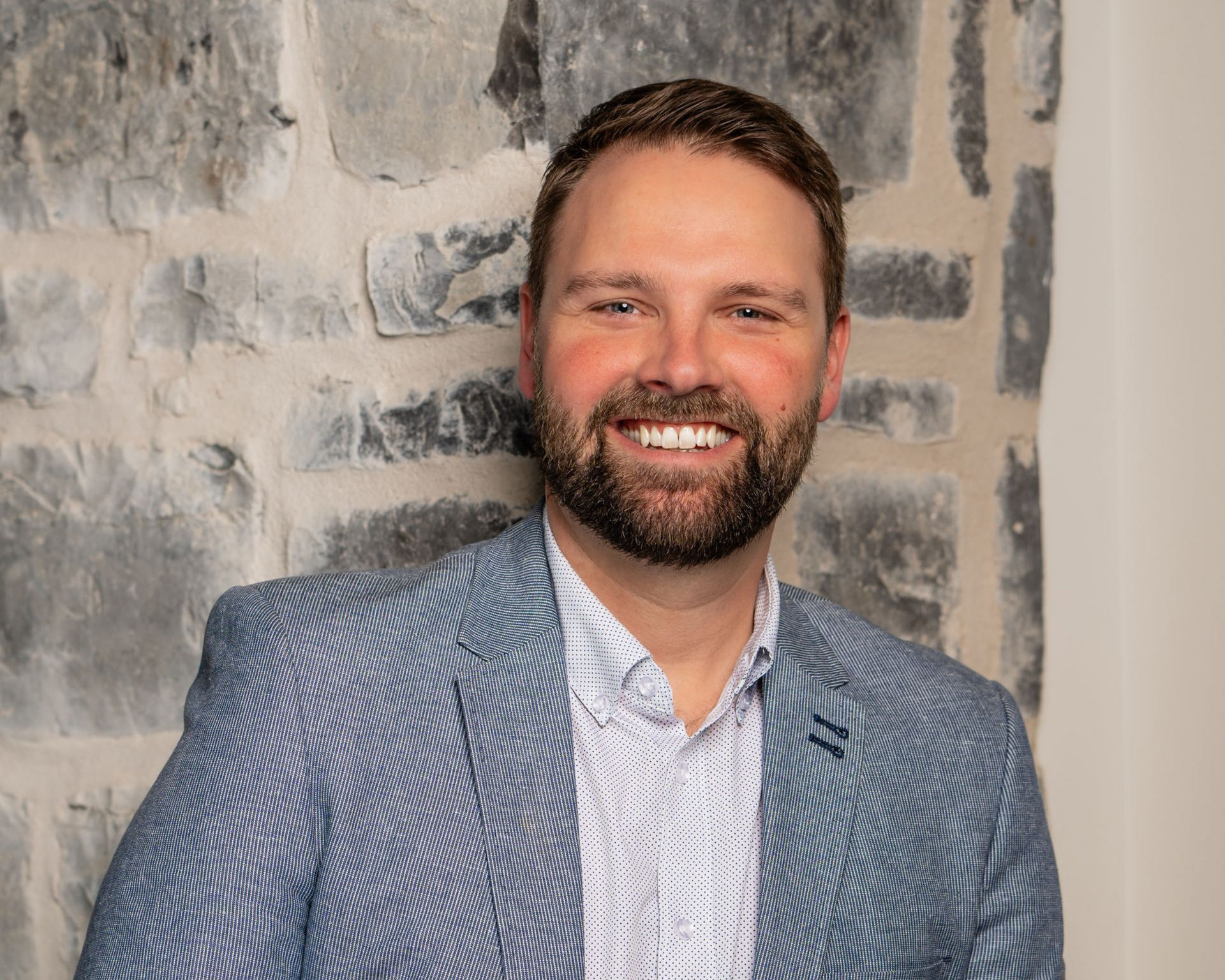 Man in blue blazer smiles, leaning against a stone wall.