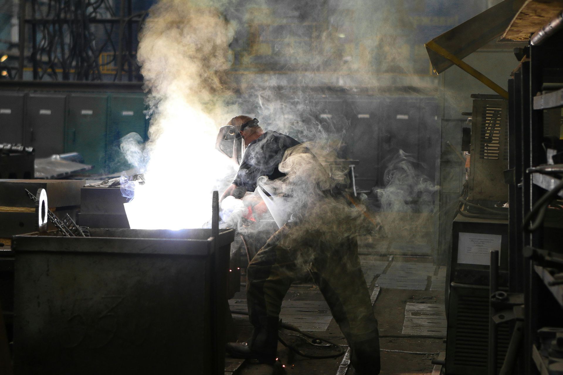 Welder in protective gear working with bright light and smoke in a workshop.