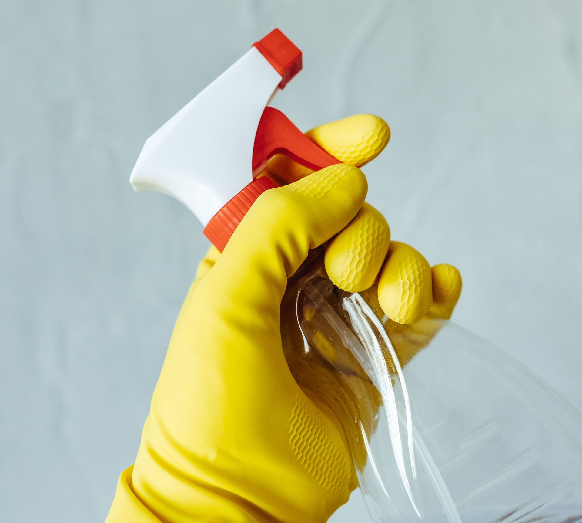 Yellow-gloved hand spraying cleaner from a white and red spray bottle into a clear glass dish