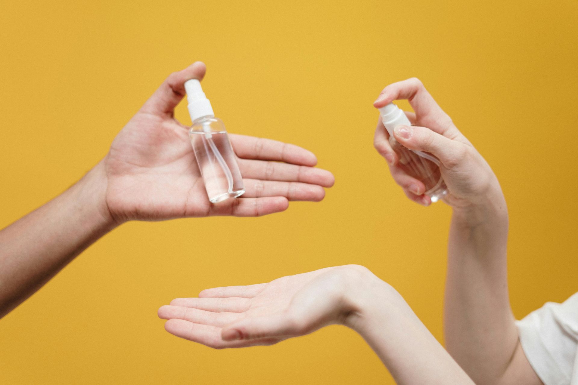 Two hands spraying sanitizer onto a third hand against a yellow background.