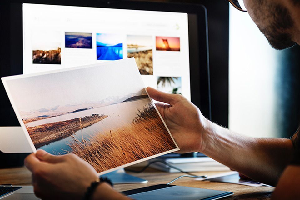 A man is holding a large printed picture of a waterfront landscape in front of a computer screen