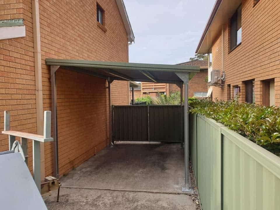 A Carport is Between Two Brick Buildings Next to a Green Fence — Hastings Valley Sheds in Wauchope, NSW