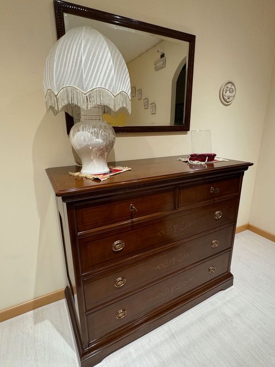 Cassettiera con lampada, specchio e decorazioni murali in una stanza. Mobili in legno scuro con ferramenta dorata.