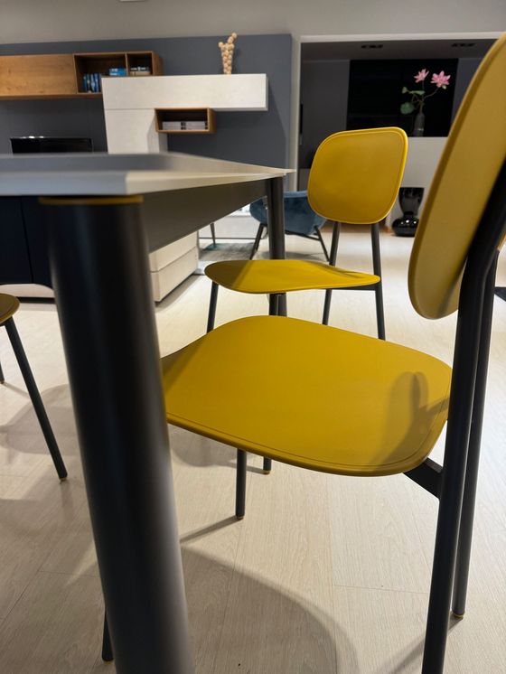 Sedie gialle circondano un tavolo in una moderna sala da pranzo. Il tavolo ha una gamba scura e sullo sfondo c'è una parete grigia.