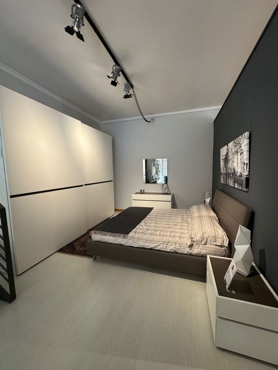 Camera da letto moderna: grande armadio bianco, letto con testiera grigia, quadro, comodino, illuminazione a binario nera.