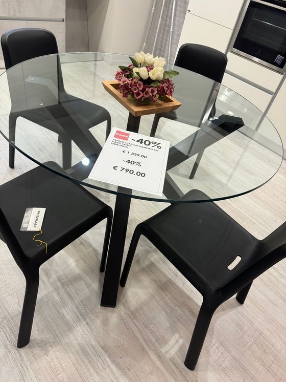 Tavolo rotondo in vetro con quattro sedie nere, fiori e cartellino del prezzo nello showroom.