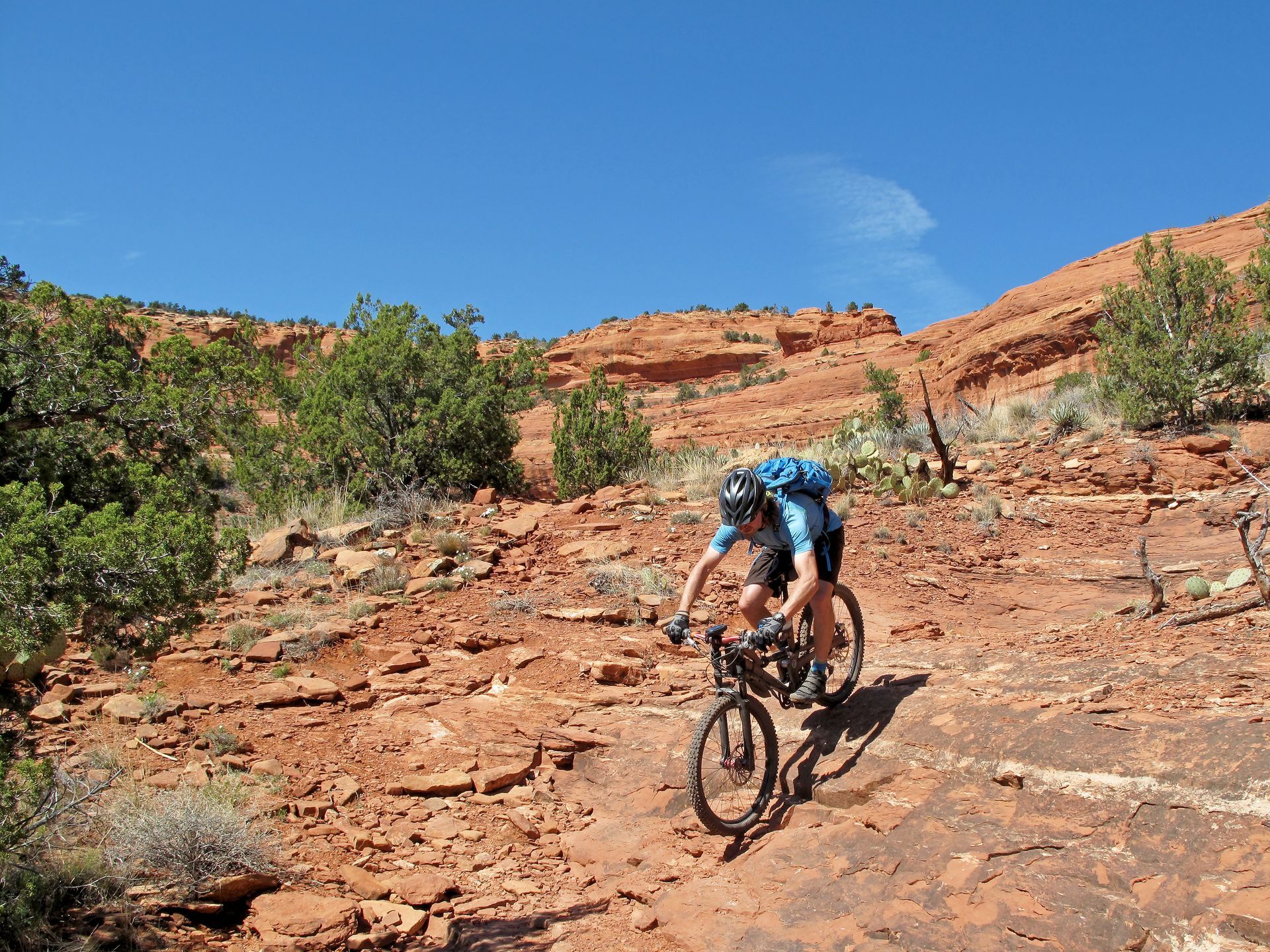 Mountain biker rides uphill on a rocky, red dirt trail in a desert landscape under a clear blue sky.