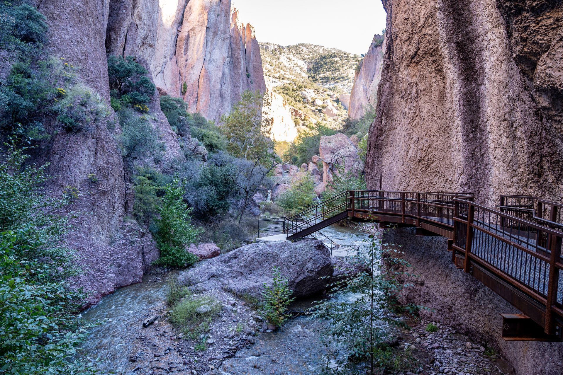 A wooden boardwalk winds through a narrow, rocky canyon with a shallow stream flowing past steep, textured cliff walls.