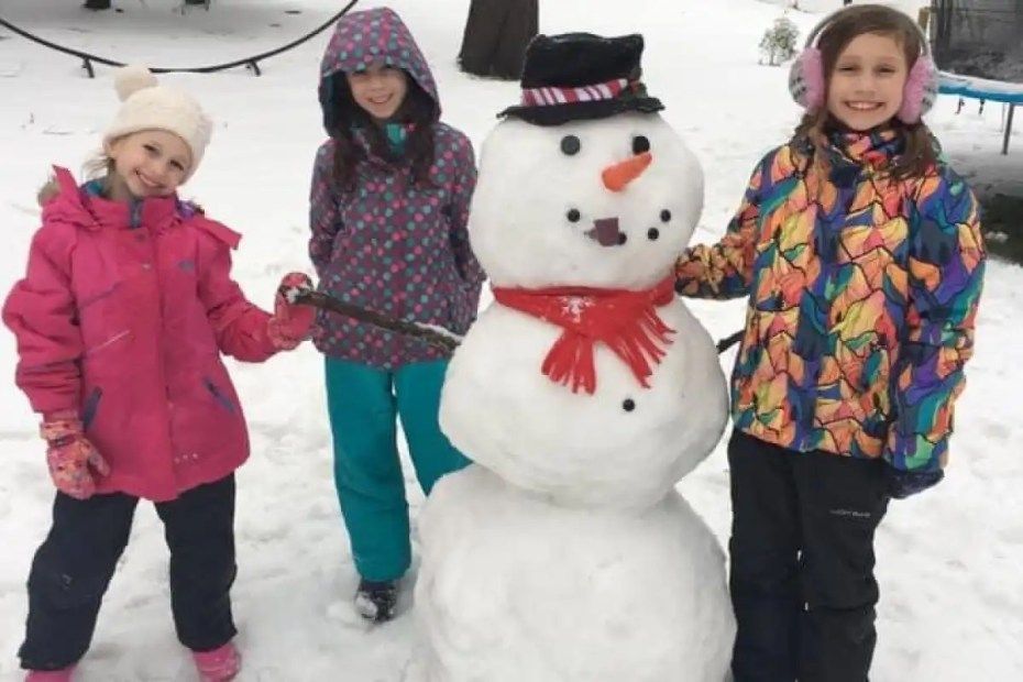 Three young girls are standing next to a snowman in the snow.