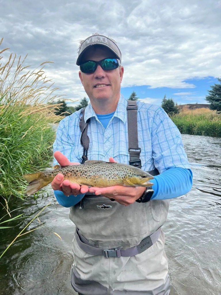 A man is holding a brown trout in his hands in front of a river.