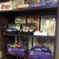 A dark brown bookshelf display holding several boxed and unboxed NASCAR toy race cars, including Jeff Gordon models.