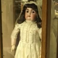 An antique doll wearing a white dress and veil, displayed inside a wooden cabinet.
