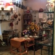 A cluttered antique shop display with wooden tables, lamps, hanging kitchen tools, and sunflowers in a vase.