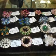 A display case filled with assorted vintage-style beaded and rhinestone brooches, each labeled with a price tag.