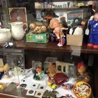 A glass display case shelf filled with vintage toys, including a tiger, dolls, a red toy car, and collectible coins.