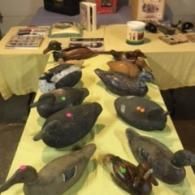 A collection of various wooden duck decoys displayed on a yellow-covered table at an indoor event.