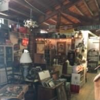 A rustic antique shop interior filled with wooden furniture, vintage decor, a white-shaded lamp, and items on shelves.