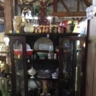 A wooden curio cabinet filled with vintage pottery, glassware, and decorative items in a rustic, indoor setting.