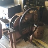 An antique, hand-cranked wooden corn sheller with a large metal flywheel stands in an indoor storage area.