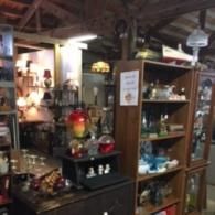 A cluttered antique shop with shelves displaying glassware, lamps, and various trinkets under exposed wooden ceiling beams.