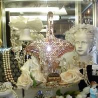 A pink glass basket sits on a display shelf surrounded by faux roses, pearl necklaces, and a small wedding cake topper.