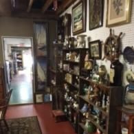 A cluttered vintage shop with shelves of collectibles, framed nautical art, and a wooden ship wheel on a wall.