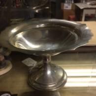 A shiny, silver-toned pedestal bowl with a wide, scalloped rim sitting on a glass table.
