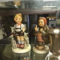 Two Hummel-style porcelain figurines, one a child with goats and the other carrying a basket, displayed behind glass.