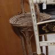 A woven wicker basket stand partially obscured by a cream-colored chair with black and gold floral-painted accents.