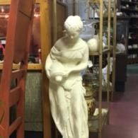 A white sculpture of a draped figure holding a vessel, standing in an antique store.