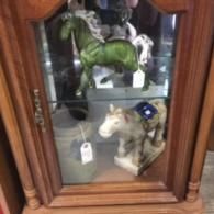A wooden display cabinet containing a green glass horse figurine on the top shelf and a grey, saddled horse on the bottom.