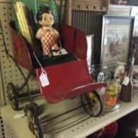 A vintage red metal doll carriage with a small, light-colored doll inside, sitting on a retail store shelf.