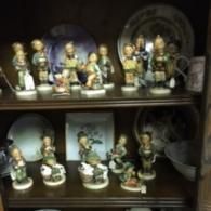 Two wooden shelves in a hutch display a collection of small Hummel-style porcelain figurines and ceramic plates.