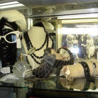 A store display case features a mannequin head with white sunglasses, beaded necklaces, vintage perfume, and jewelry.