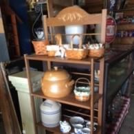 A wooden tiered display shelf in a shop, holding various woven baskets, a large tan ceramic pot, a stoneware crock, and mugs.