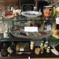 A multi-tiered glass display shelf filled with various vintage trinkets, figurines, a clear ashtray, and a tiki-style mug.