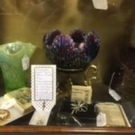 A display case with a purple iridescent glass bowl, a green glass vase, antique books, and a metallic brooch.