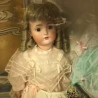 An antique porcelain doll with curly hair and a white lace dress displayed inside a glass case.