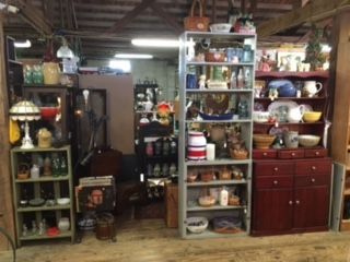 Shelves filled with various antiques, glassware, and home decor items inside a cluttered, wood-beamed vintage store.