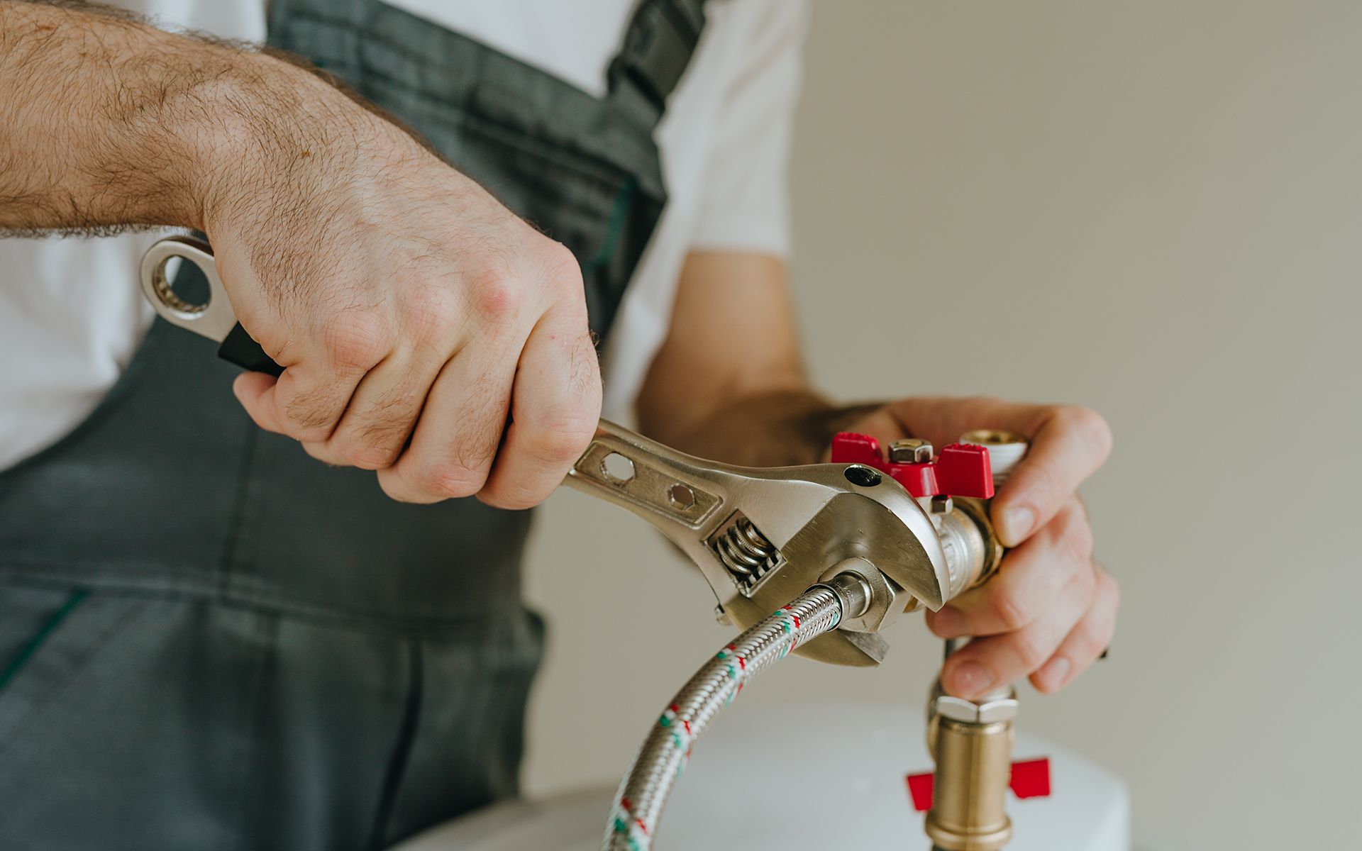 Plumber in gray overalls using a wrench on a pipe with red valves.