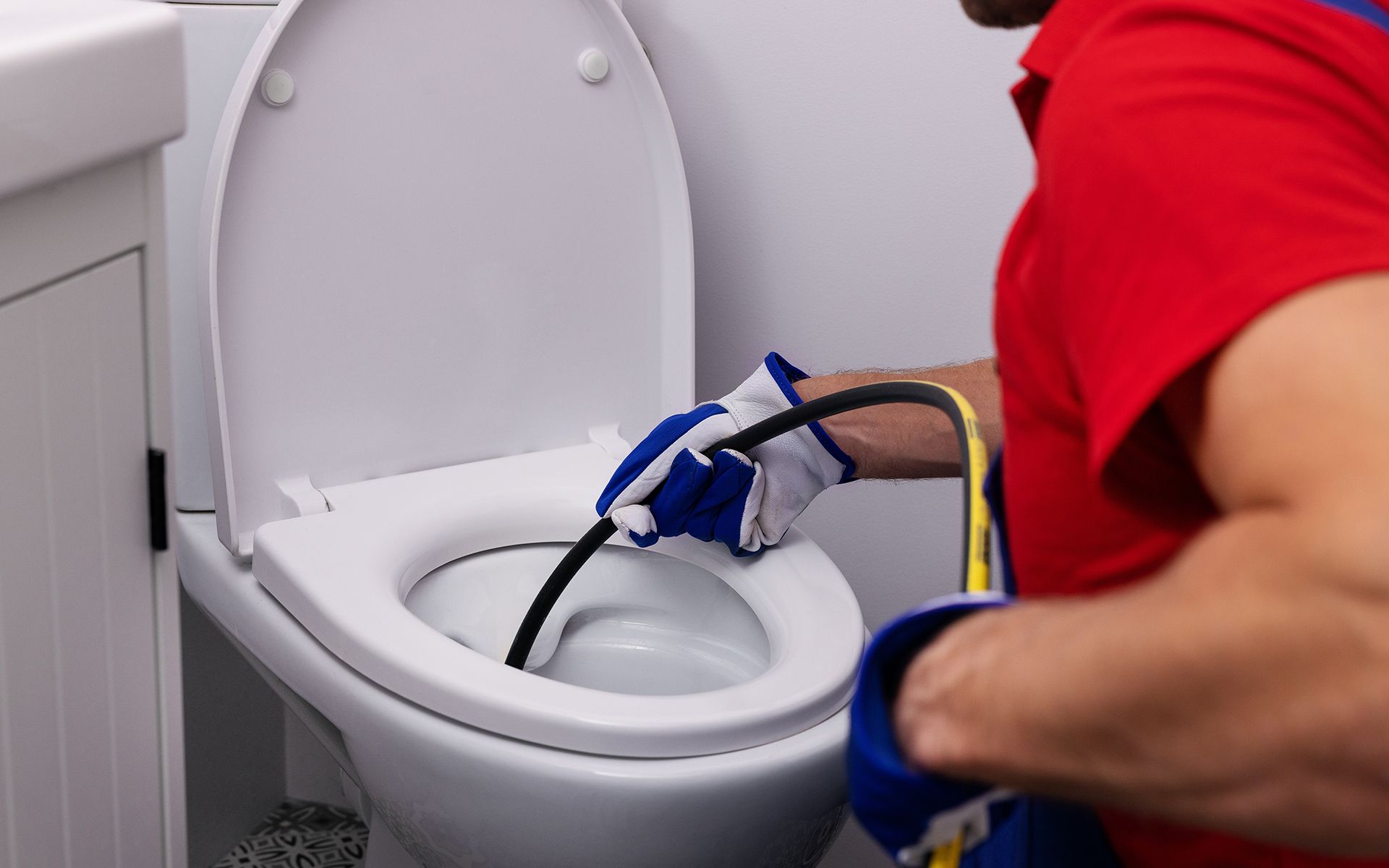 Plumber in red shirt and gloves unclogging a white toilet with a snake in a bathroom.