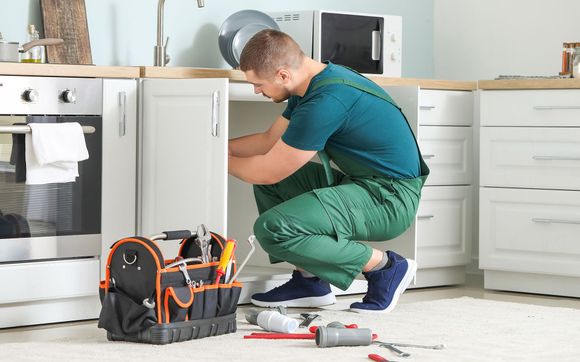 Man in green jumpsuit, crouching inside a kitchen cabinet, working. Tools in a toolbox and on the floor.