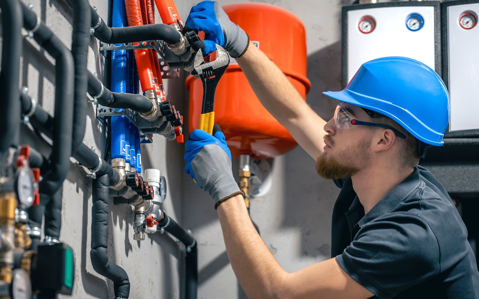 Plumber in blue helmet and gloves, using a wrench on pipes in an industrial setting.