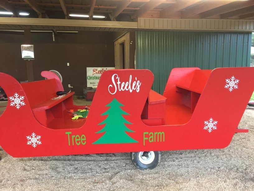 Red sleigh on wheels, Steele's Tree Farm. Green tree graphic, white snowflakes, under awning with green and brown wall.